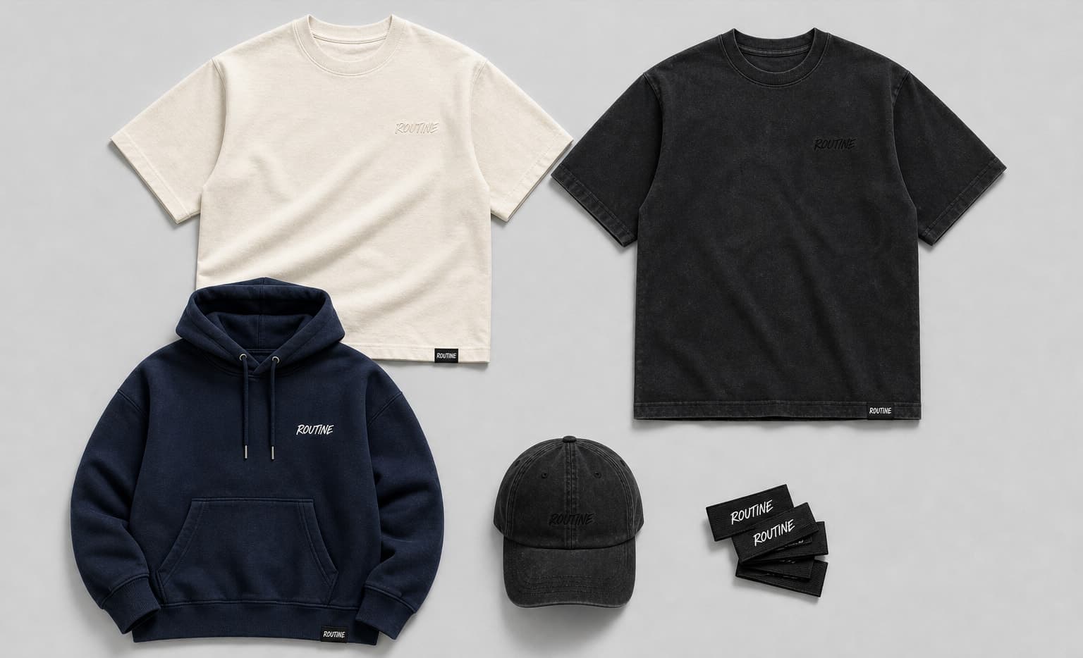 Routine Drop 001 Essentials board. Cream heavyweight tee, washed-black heavyweight tee, navy fleece hoodie and washed-black dad hat, each with a small embroidered ROUTINE script and a woven hem tag.