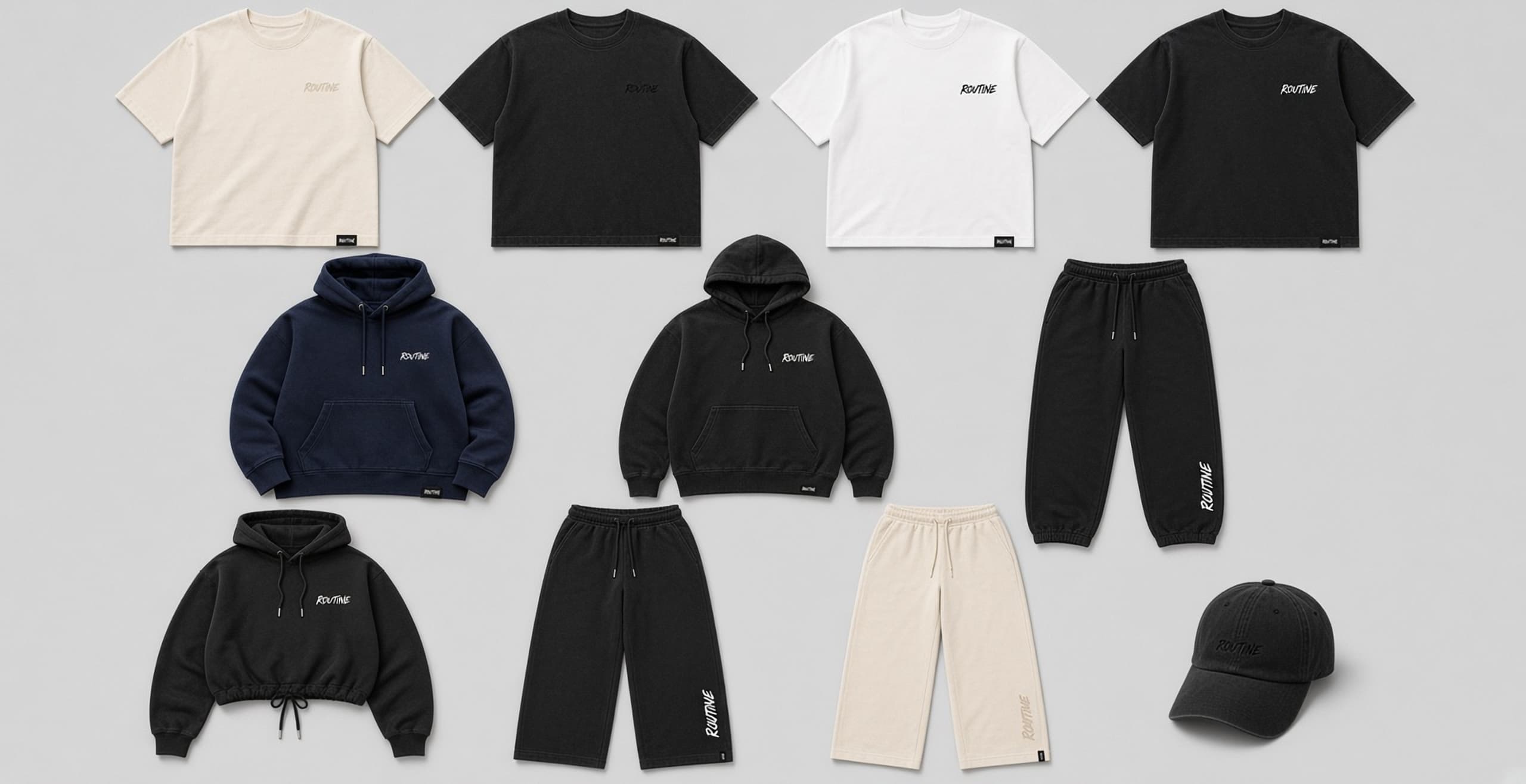 Flat lay of Essentials Drop 001 garments with small embroidered ROUTINE script and woven hem tags. Cream, washed black, white, and navy pieces on a clean surface.