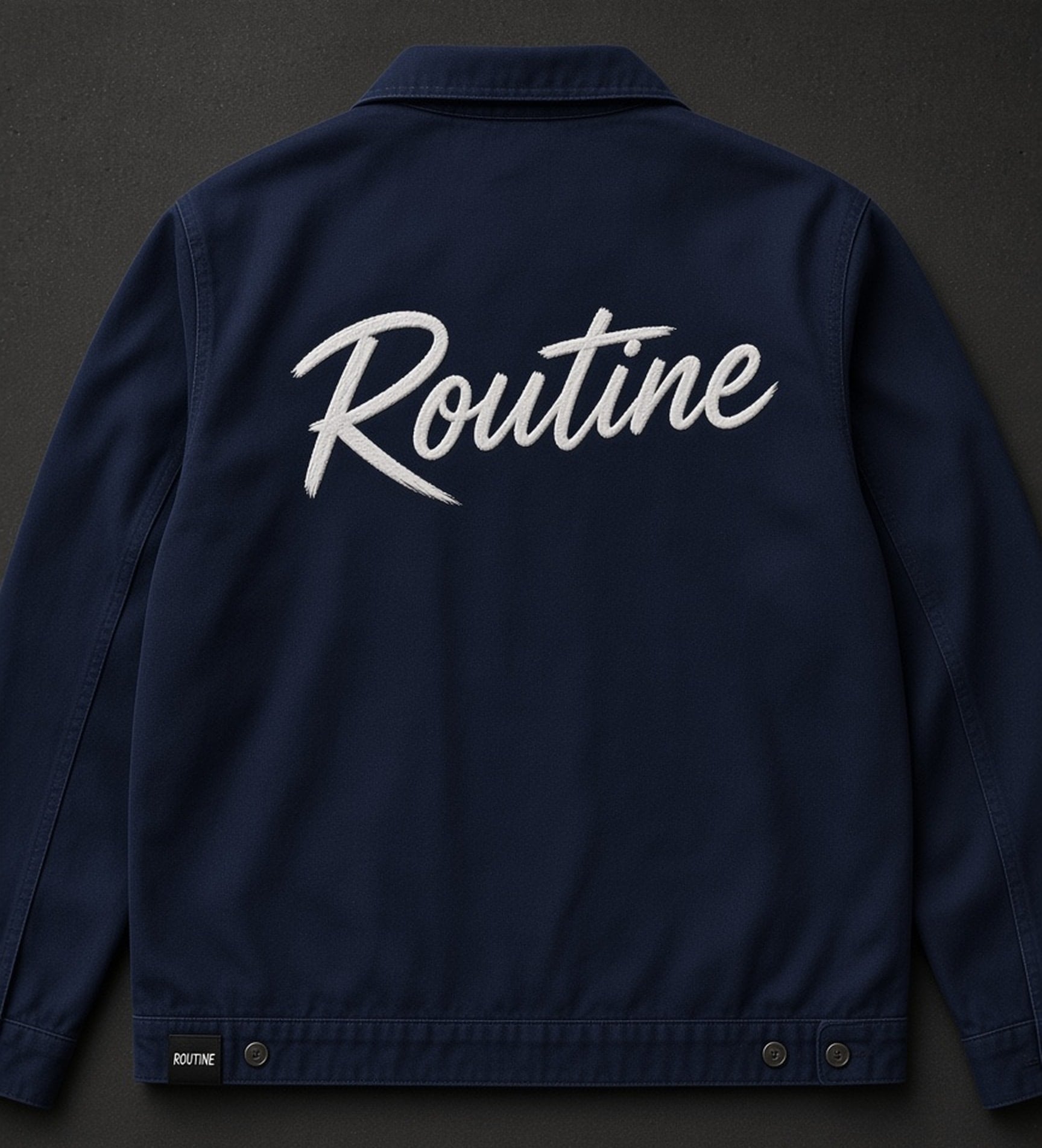 Navy work jacket laid flat, back view. Large white brush script ROUTINE across the upper back and a black ROUTINE hem tag at the left hem.